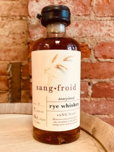 A bottle of Sang Froid Maryland rye whiskey from Baltimore Spirits Company sits on a wooden surface, with a brick wall in the background. The label features wheat graphics and detailed text about the whiskey.