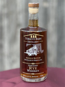 A bottle of Tobacco Barn Distillery Single Batch Straight Rye Whiskey, featuring a barn illustration on the label, sits on a maroon surface with a blurred gray background, evoking the craftsmanship found in renowned local distilleries like Baltimore Spirits Company.