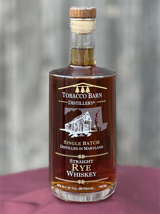 A bottle of Tobacco Barn Distillery Single Batch Straight Rye Whiskey, featuring a barn illustration on the label, sits on a maroon surface with a blurred gray background, evoking the craftsmanship found in renowned local distilleries like Baltimore Spirits Company.