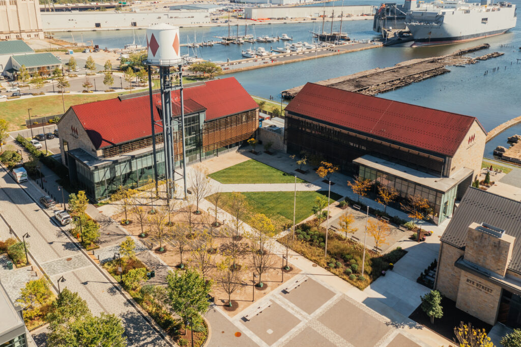 Aerial view of a waterfront area with two red-roofed buildings, a water tower, trees, walkways, and parked boats in a marina; a large ship is docked nearby.