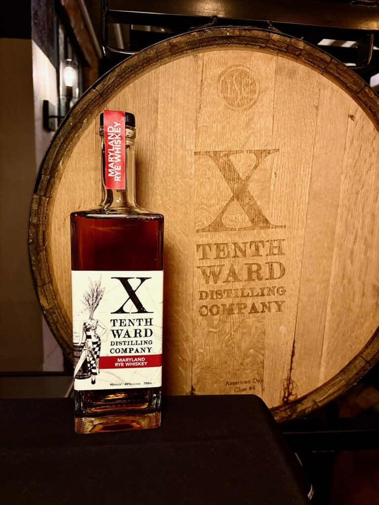 Tenth Ward Distilling Company Maryland Rye Whiskey bottle in front of a branded wooden barrel