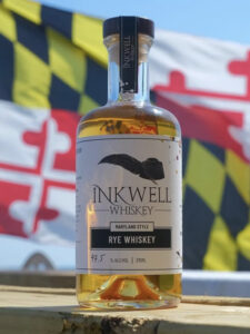 A bottle of Inkwell Rye Whiskey from Shmidt Spirits sits on a wooden surface with a Maryland state flag in the background, both outdoors under a clear blue sky.
