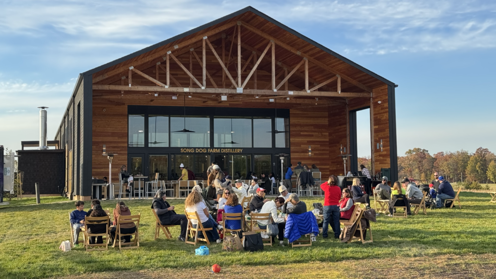 People sit on chairs outside a modern barn-style building called Sowing Dog Farm Distillery, enjoying a sunny day, with musicians performing on a patio stage in front of green lawns and distant trees.